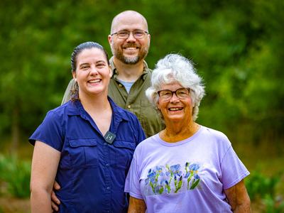 Three adults standing outdoors; front woman wearing light purple shirt with floral print.