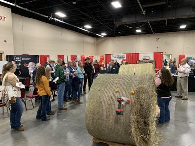 People inspecting large round hay bales at an indoor agricultural trade show