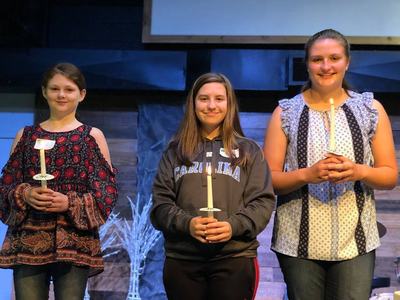 Three girls standing on stage holding lit candles; center wearing "CAROLINA" sweatshirt