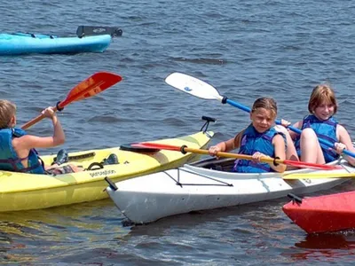 Three kids wearing life jackets paddling in yellow, white, and red kayaks on water