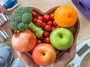 Hands holding heart-shaped bowl of fruit and vegetables; dumbbells, scale, yoga mat nearby