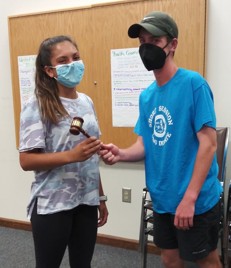 Two masked youths indoors, one handing a wooden gavel to the other; poster says "Youth Council."