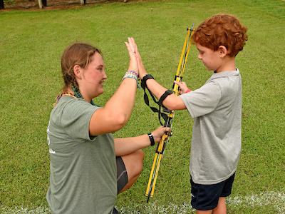 Adult instructor kneeling gives high-five to child holding a youth bow at archery range