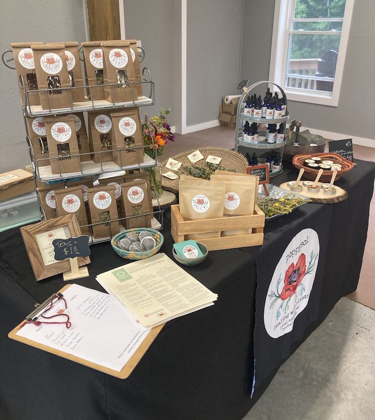 Vendor table displaying packaged teas, tinctures, samples, sign "Teas $13" and logo