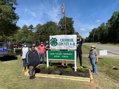 Group of masked volunteers holding "CHATHAM COUNTY 4-H" banner by community action center sign