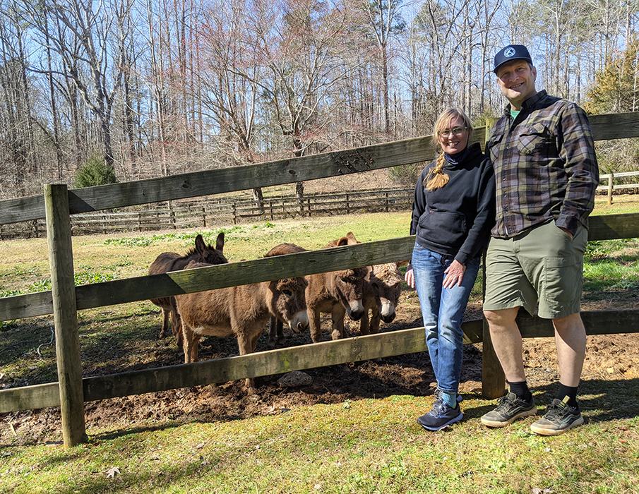 Two people standing by wooden fence with three brown donkeys in pasture