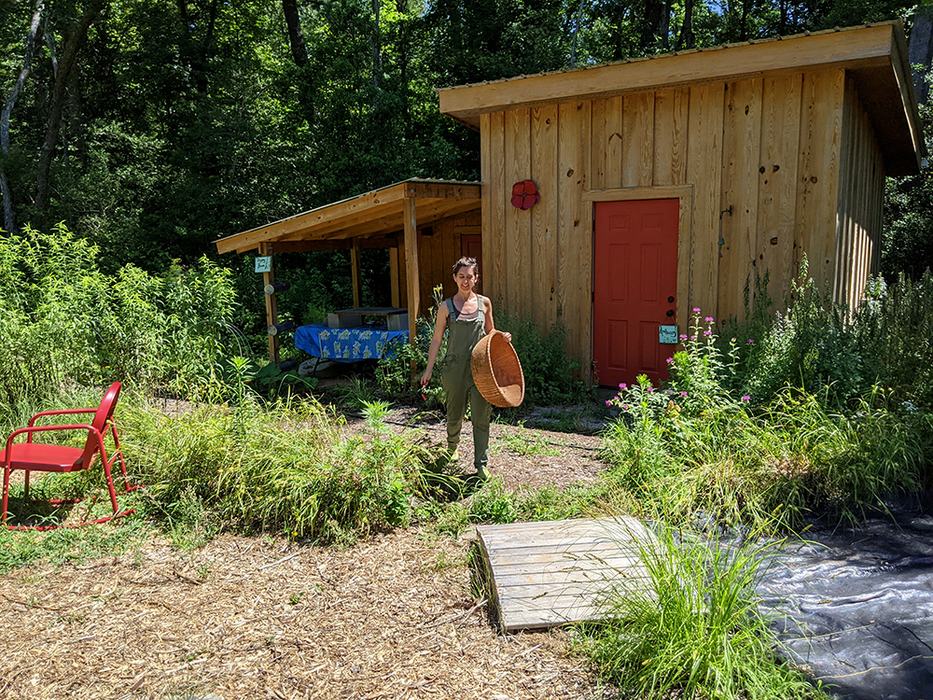 Person in green overalls holding a large wicker basket standing in front of a wooden shed