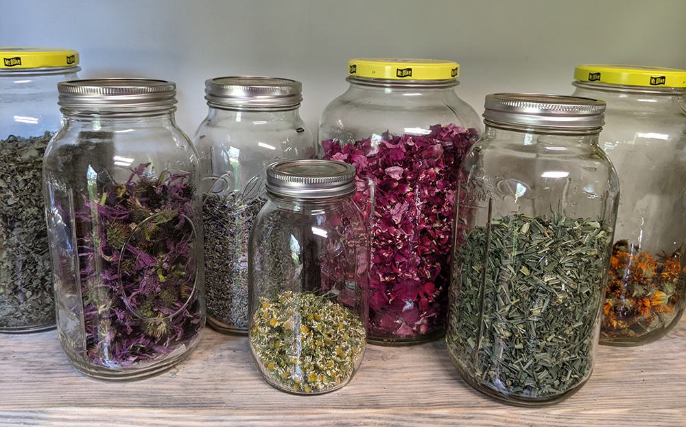 Glass jars on a shelf holding assorted dried herbs and flower petals