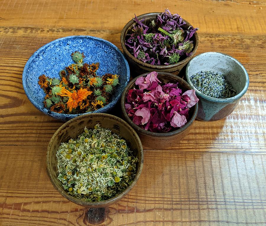 Five bowls of dried herbs and flower petals on a wooden table