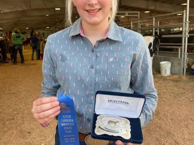 Person holding blue ribbon "North Carolina Mountains State Fair" and boxed "Montana" belt buckle in barn
