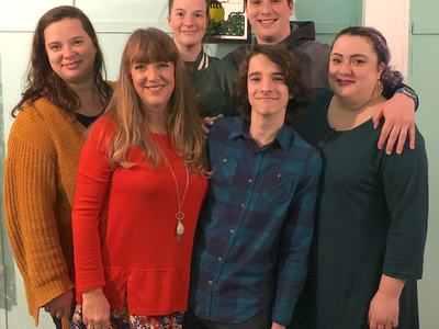 Six people standing close together indoors; framed sign behind reads "To make the best"