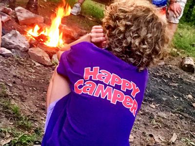 Person with curly hair sitting by a campfire wearing a purple shirt reading "HAPPY CAMPER"