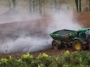 Green tractor spreading granular fertilizer across plowed field, white dust cloud