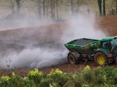 Green tractor spreading granular fertilizer across plowed field, white dust cloud