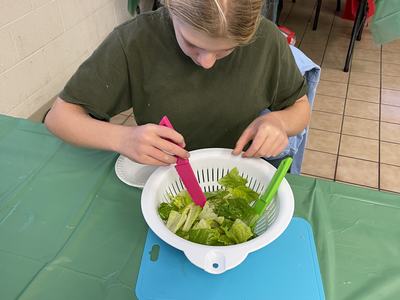 Child mixing romaine lettuce in a white colander using pink and green tongs at a table