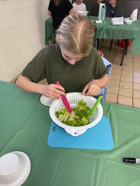 Child mixing romaine lettuce in a white colander using pink and green tongs at a table