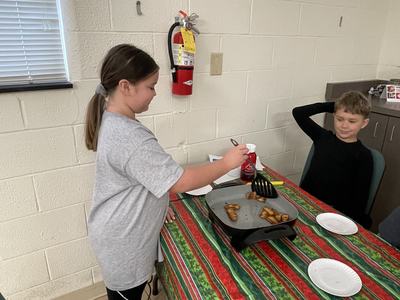 Girl using spatula to cook potato pieces on electric griddle at table; boy seated
