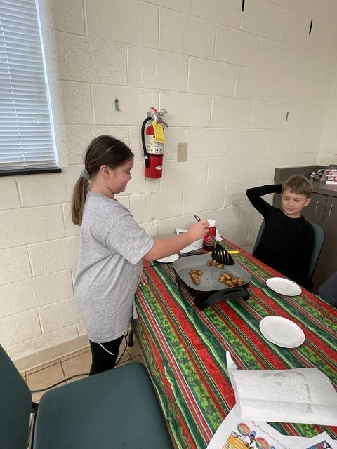 Girl using spatula to cook potato pieces on electric griddle at table; boy seated