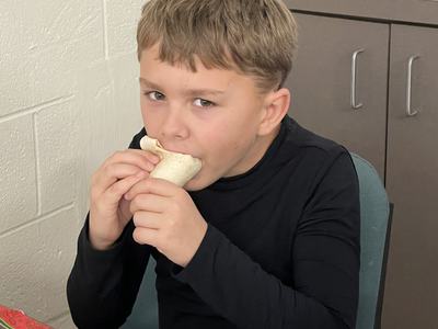 Young boy biting into a wrapped tortilla/burrito while seated at a table with a paper plate.