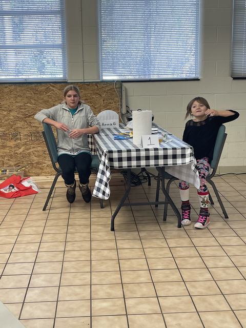 Two children seated at a checkered table with paper towel, sign "1", and paper labeled "Anna :)"