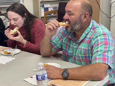 Man in plaid shirt biting a pastry at a table while a woman beside him also eats