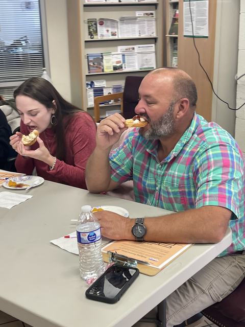 Man in plaid shirt biting a pastry at a table while a woman beside him also eats
