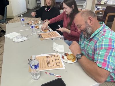 Three people seated at a table eating from plates with water bottles and clipboards