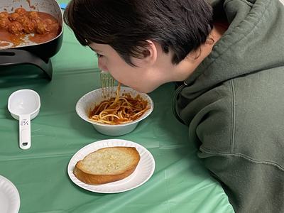 Person leaning over a bowl, eating spaghetti with a fork; garlic bread and meatballs nearby