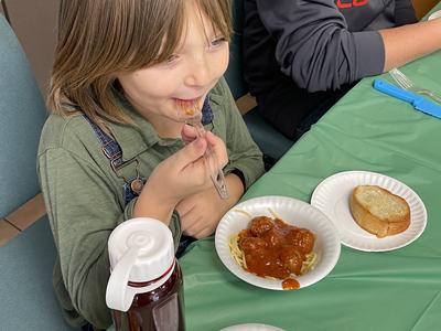Child eating spaghetti and meatballs with bread and water bottle at green-covered table