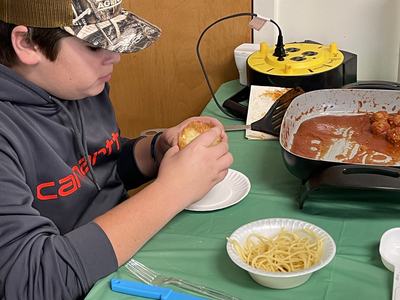 Person in Carhartt hoodie holding a roll beside a bowl of spaghetti and a pan of meatballs