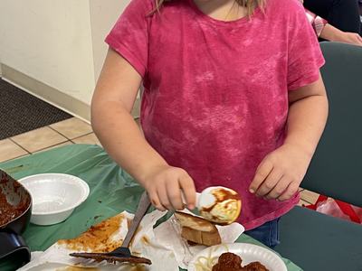 Girl in pink shirt spooning sauce onto bread near bowl of spaghetti and meatballs.
