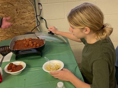 Person lifting lid off a pan of meatballs while holding a bowl of spaghetti at a table