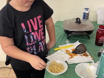 Child serving spaghetti from a bowl at a table with skillet and bread; shirt reads "LIVE LOVE PLAY"