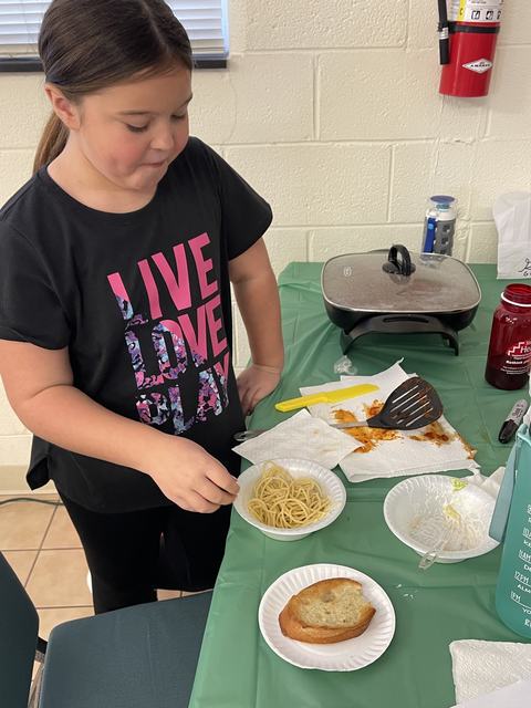 Child serving spaghetti from a bowl at a table with skillet and bread; shirt reads "LIVE LOVE PLAY"