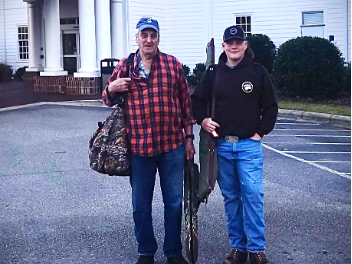 Two people by white-columned building; older man with camo bag, younger holding camo gear