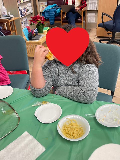 Child sitting at table eating a bread roll, bowls of spaghetti and paper plates on green tablecloth