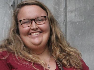 Person wearing glasses and red shirt, long blond hair, cross necklace against gray wall