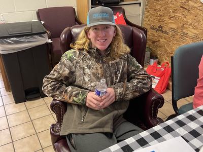 Person in camo jacket and "SIMPLY NATURAL CREAMERY" hat sitting in armchair holding a water bottle