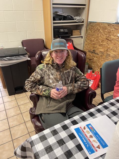 Person in camo jacket and "SIMPLY NATURAL CREAMERY" hat sitting in armchair holding a water bottle
