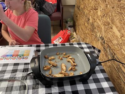 Electric skillet with cooked chicken pieces on checkered tablecloth; girl drinking from bottle in background.