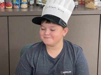 Child wearing paper chef hat labeled "Jackson King," seated at table with canned food behind