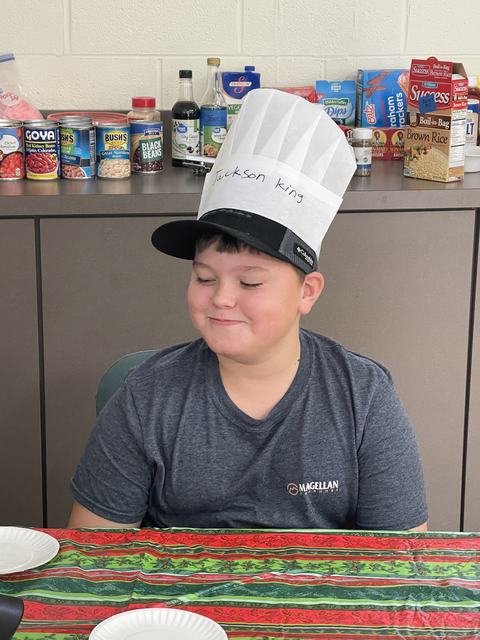 Child wearing paper chef hat labeled "Jackson King," seated at table with canned food behind