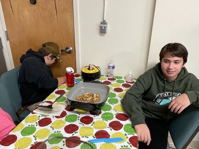 Two boys sitting at a table with an electric skillet cooking chicken on apple-pattern tablecloth