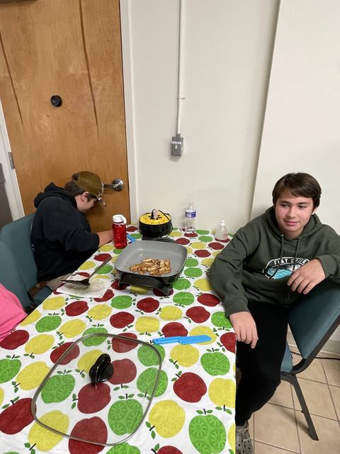 Two boys sitting at a table with an electric skillet cooking chicken on apple-pattern tablecloth