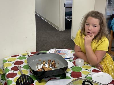 Girl in yellow seated at table beside electric skillet with small cooked meat pieces