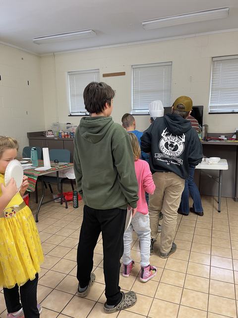 Group of people lined up at a serving table in a community room; hoodie reads "Eastern Outfitters"