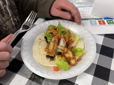 Person holding fork over chicken taco with lettuce, tomato, and black beans on paper plate