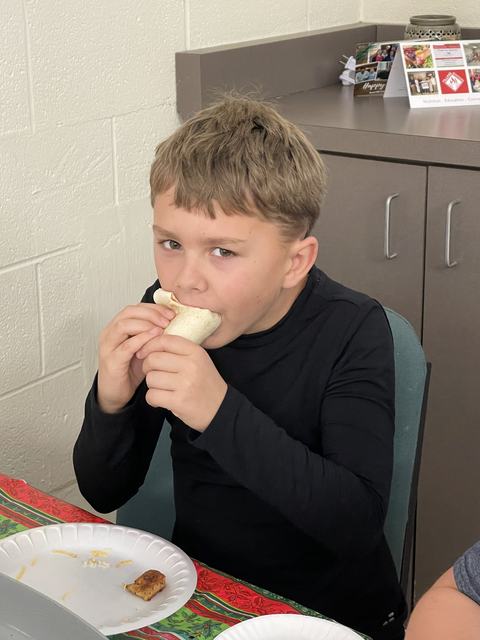 Young boy biting into a wrapped tortilla at a table with paper plates.