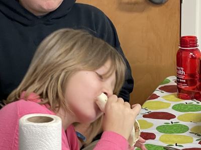 Girl in pink shirt biting a tortilla wrap at a table, person in hat seated behind