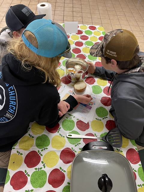 Three children in caps leaning over table with a metal bowl of condiments and a bread roll